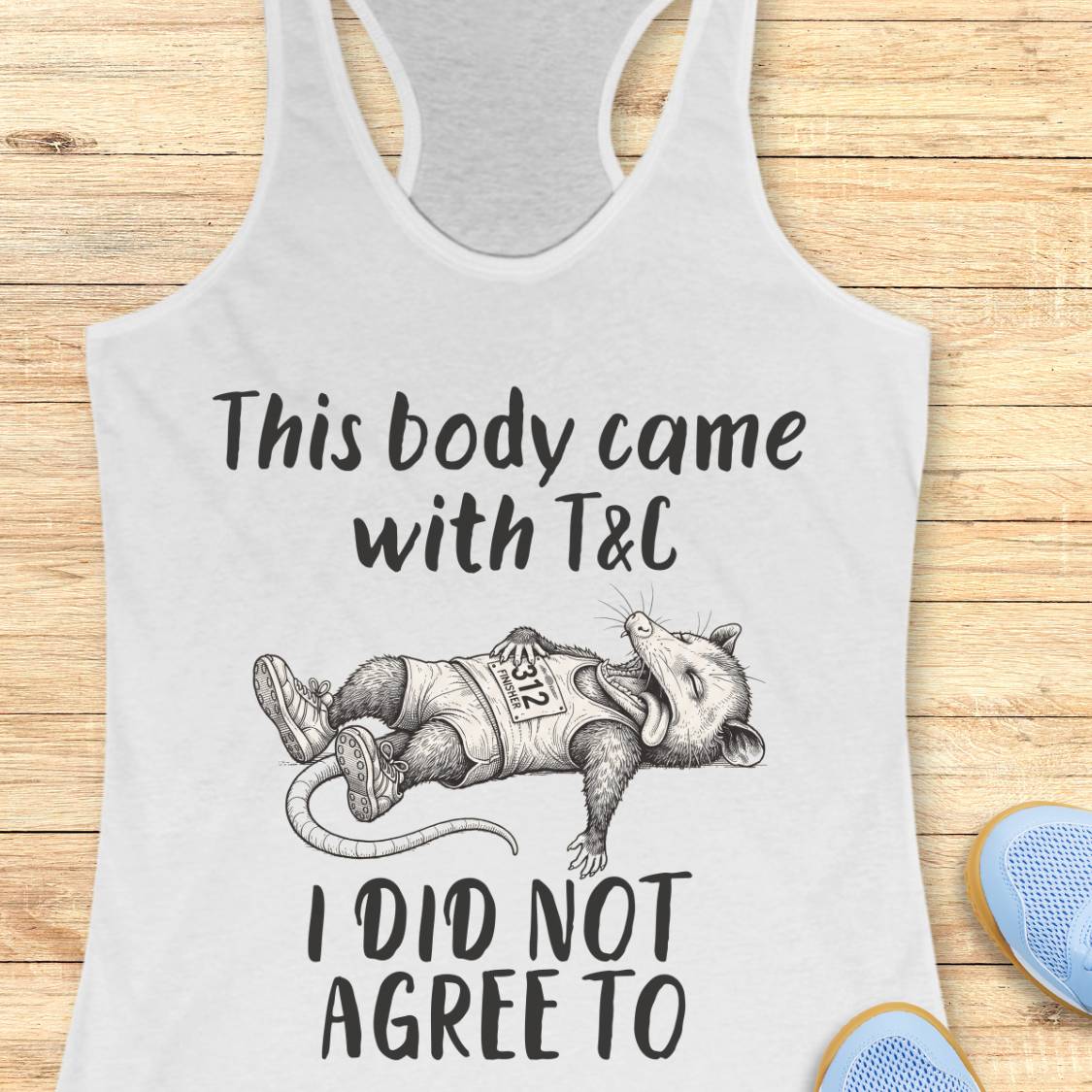 A white racerback tank top laid flat on a light wood background, featuring a black graphic of a sleeping raccoon wearing a race bib and the text 'This body came with T&C I DID NOT AGREE TO'. Partial blue shoes are visible at the bottom right as styling props.