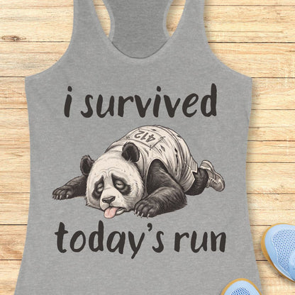 A heather gray racerback tank top laid flat on a light wood surface, featuring a black graphic of a panda wearing a race bib and the text 'i survived today's run'. Partial blue shoes are visible at the bottom right edge.