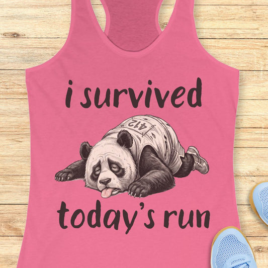 A pink racerback tank top laid flat on a light wood surface, featuring a black graphic of a panda wearing a race jersey with the text 'i survived today's run'. Partial blue sneakers are visible at the bottom right as styling props.