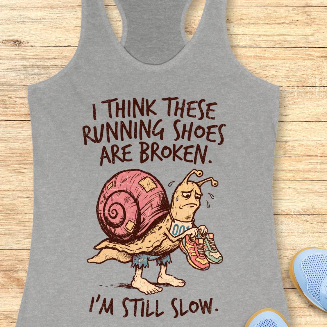 A heather gray racerback tank top laid flat on a light wood surface, featuring a cartoon snail wearing pink running shoes and the text 'I THINK THESE RUNNING SHOES ARE BROKEN. I'M STILL SLOW.
