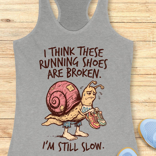 A heather gray racerback tank top laid flat on a light wood surface, featuring a cartoon snail wearing pink running shoes and the text 'I THINK THESE RUNNING SHOES ARE BROKEN. I'M STILL SLOW.