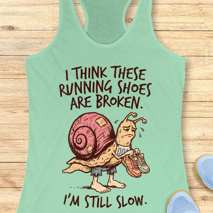 A mint green racerback tank top laid flat on a light wood background, featuring a cartoon snail graphic and the text 'I THINK THESE RUNNING SHOES ARE BROKEN. I'M STILL SLOW.