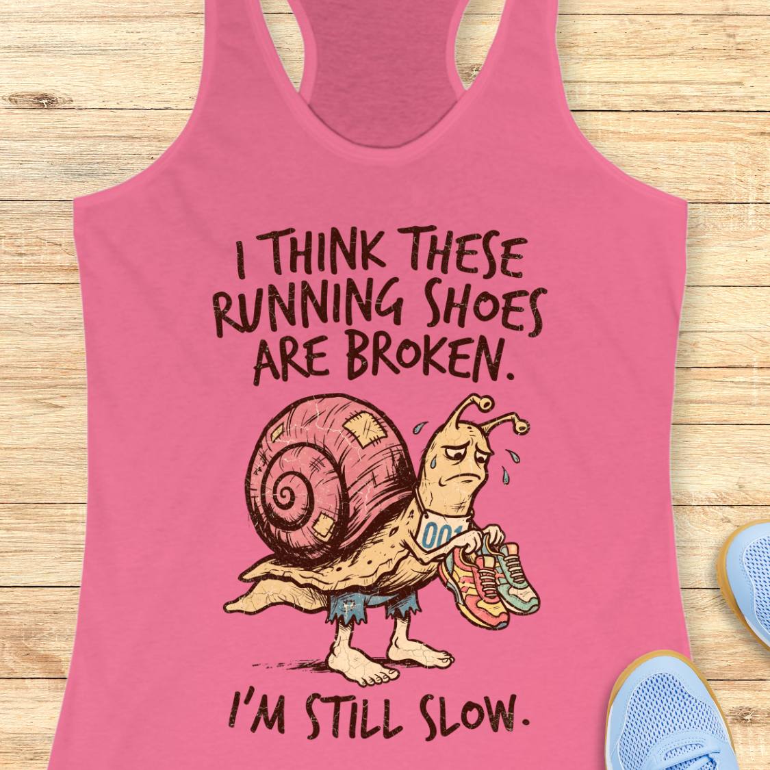 A pink racerback tank top laid flat on a light wood surface, featuring a cartoon snail holding broken running shoes with the text 'I THINK THESE RUNNING SHOES ARE BROKEN. I'M STILL SLOW.