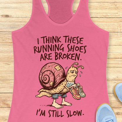 A pink racerback tank top laid flat on a light wood surface, featuring a cartoon snail holding broken running shoes with the text 'I THINK THESE RUNNING SHOES ARE BROKEN. I'M STILL SLOW.