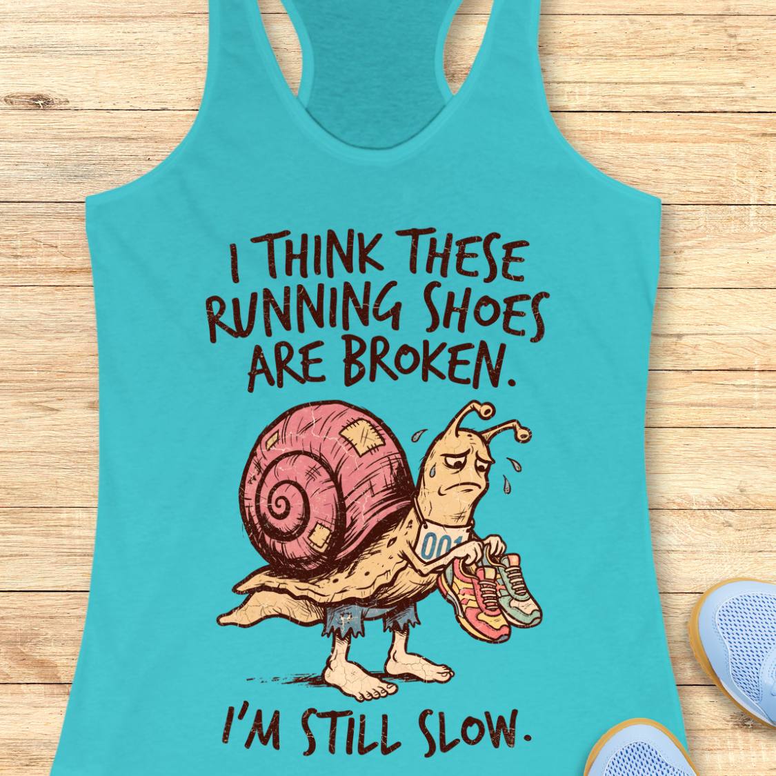 A turquoise racerback tank top laid flat on a light wood surface, featuring a cartoon snail wearing broken running shoes with the text 'I THINK THESE RUNNING SHOES ARE BROKEN. I'M STILL SLOW.