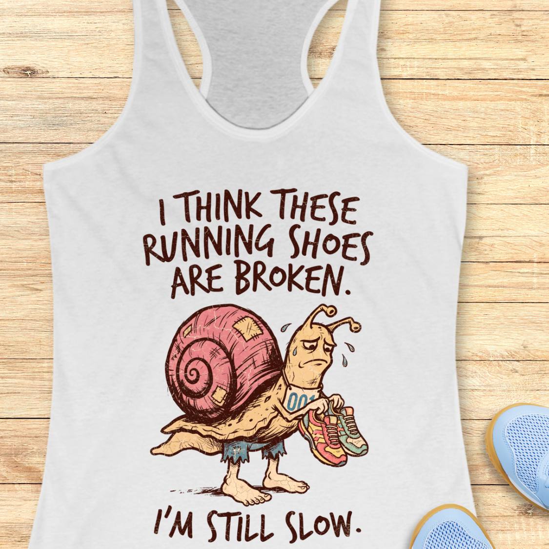 A white racerback tank top laid flat on a light wood surface, featuring a cartoon snail carrying running shoes with the text 'I THINK THESE RUNNING SHOES ARE BROKEN. I'M STILL SLOW.
