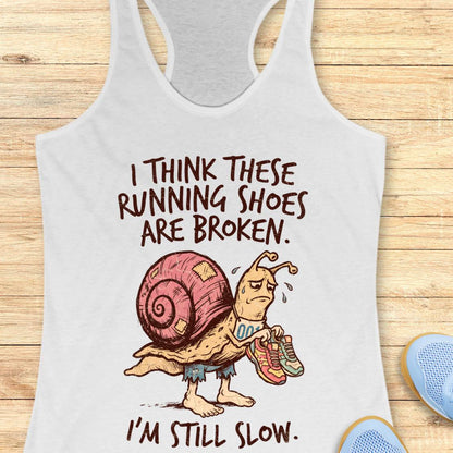 A white racerback tank top laid flat on a light wood surface, featuring a cartoon snail carrying running shoes with the text 'I THINK THESE RUNNING SHOES ARE BROKEN. I'M STILL SLOW.