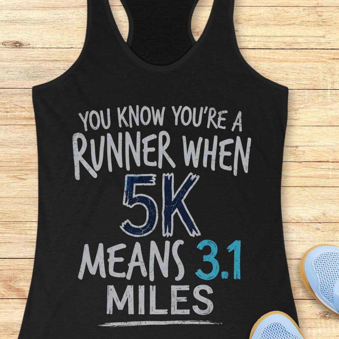 A black racerback tank top laid flat on a light wood surface, printed with the phrase 'YOU KNOW YOU'RE A RUNNER WHEN 5K MEANS 3.1 MILES' in white, blue, and teal lettering.