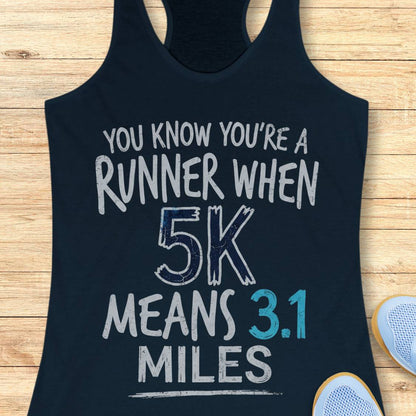 A navy racerback tank top laid flat on a light wood surface, printed with the phrase 'YOU KNOW YOU'RE A RUNNER WHEN 5K MEANS 3.1 MILES' in distressed white and blue lettering.