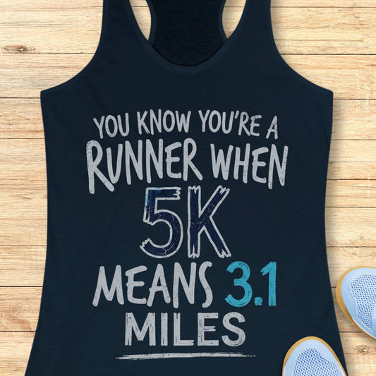 A navy racerback tank top laid flat on a light wood surface, printed with the phrase 'YOU KNOW YOU'RE A RUNNER WHEN 5K MEANS 3.1 MILES' in distressed white and blue lettering.