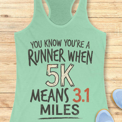 A mint green racerback tank top with a humorous 5K running slogan printed on the front, laid flat on a light wood background with a pair of blue sneakers partially visible in the corner.