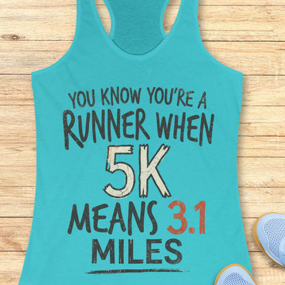 A turquoise racerback tank top laid flat on a light wood surface, printed with the phrase 'YOU KNOW YOU'RE A RUNNER WHEN 5K MEANS 3.1 MILES' in black, white, and orange text.