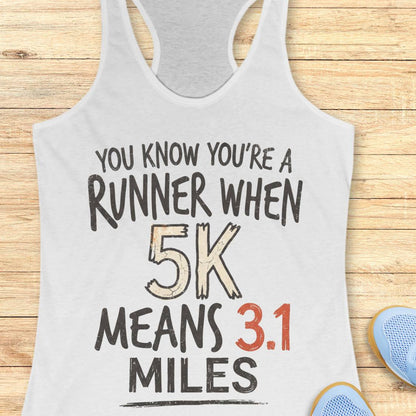 A white racerback tank top laid flat on a light wood background, printed with the phrase 'YOU KNOW YOU'RE A RUNNER WHEN 5K MEANS 3.1 MILES' in black and red lettering.