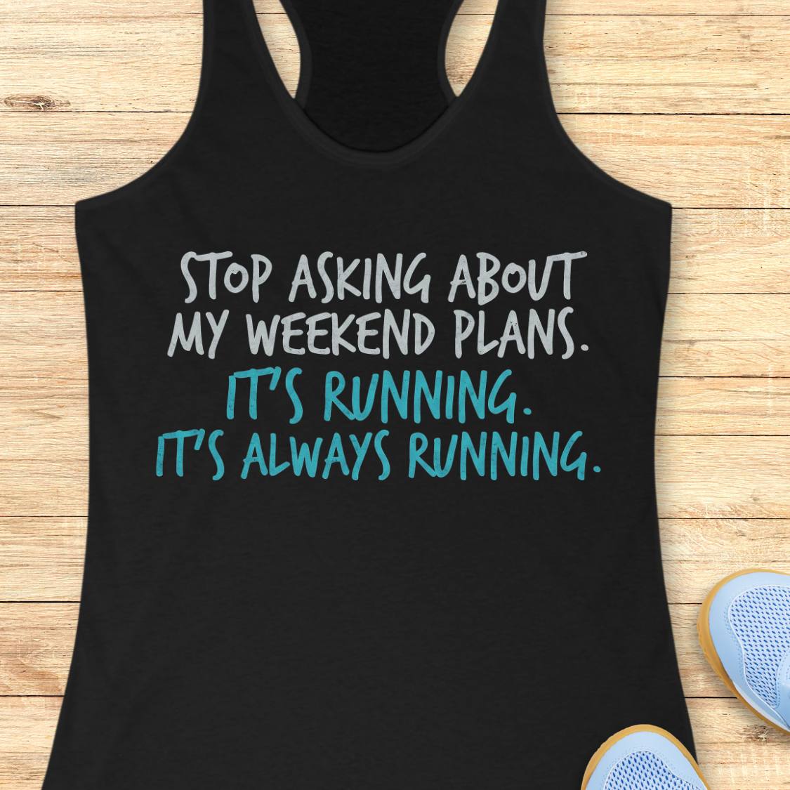 A black racerback tank top laid flat on a light wood surface, printed with the phrase 'STOP ASKING ABOUT MY WEEKEND PLANS. IT'S RUNNING.