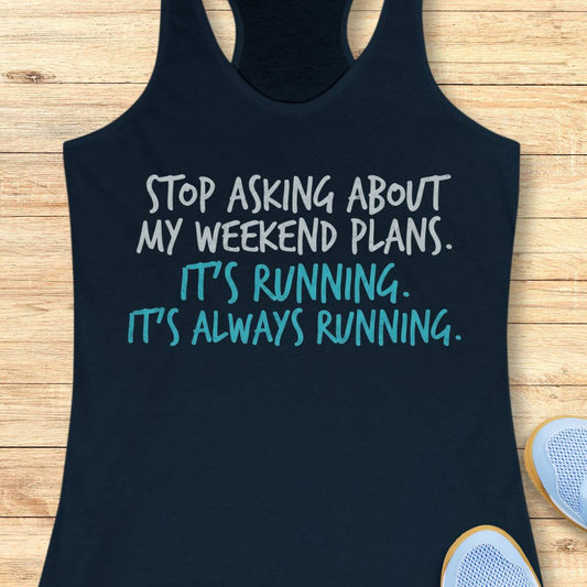 A navy racerback tank top laid flat on a light wood surface, printed with the phrase 'STOP ASKING ABOUT MY WEEKEND PLANS. IT'S RUNNING.