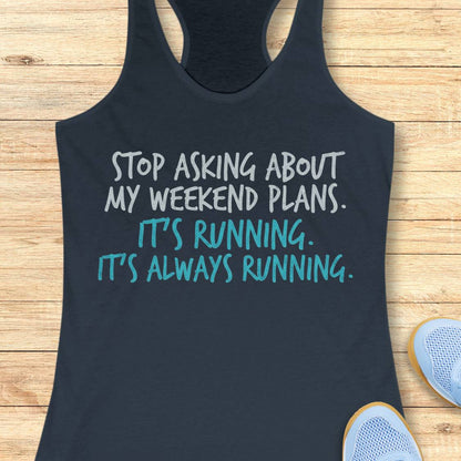 A indigo racerback tank top laid flat on a light wood background, with white and teal text that reads, 'STOP ASKING ABOUT MY WEEKEND PLANS. IT'S RUNNING.