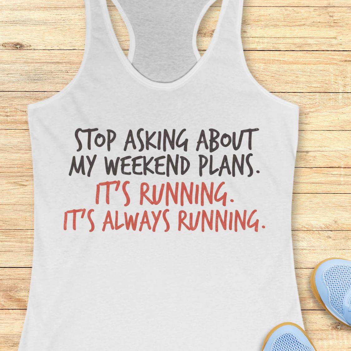 A white racerback tank top laid flat on a light wood plank background, with printed text reading 'STOP ASKING ABOUT MY WEEKEND PLANS. IT'S RUNNING.