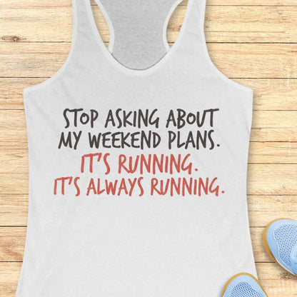 A white racerback tank top laid flat on a light wood plank background, with printed text reading 'STOP ASKING ABOUT MY WEEKEND PLANS. IT'S RUNNING.