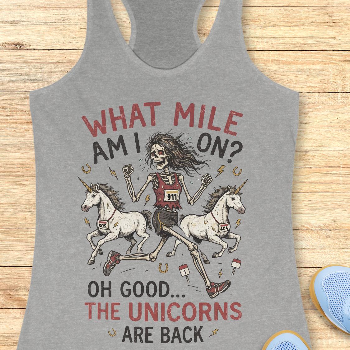 A heather gray racerback tank top laid flat on a light wood background, featuring a graphic of a skeleton runner riding two white unicorns with the text 'WHAT MILE AM I ON? OH GOOD.