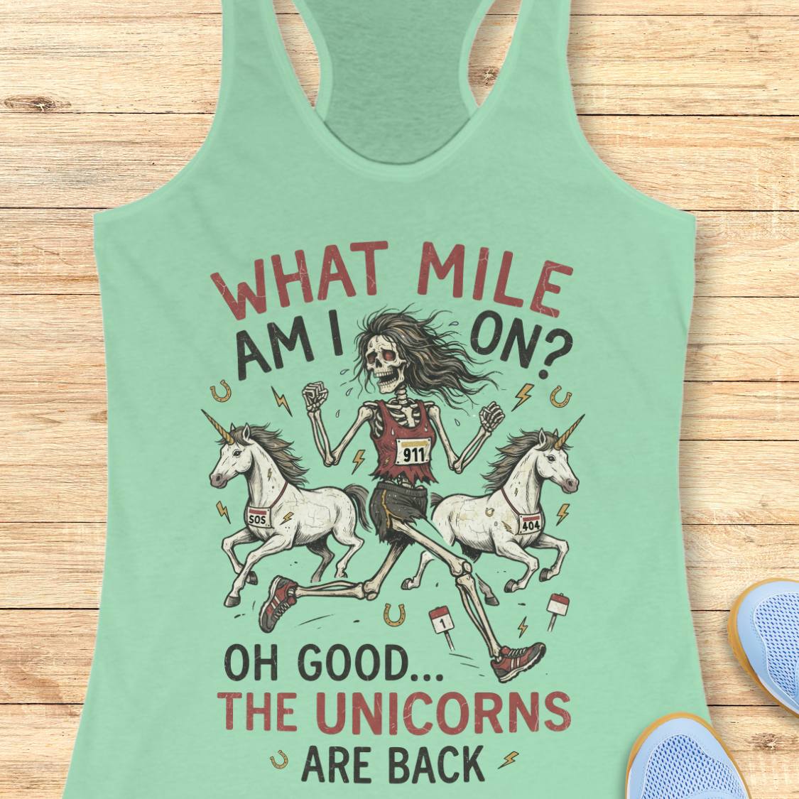 A mint green racerback tank top laid flat on a light wood background, featuring a humorous graphic of a skeleton race runner with two white unicorns and the text 'WHAT MILE AM I ON? OH GOOD.