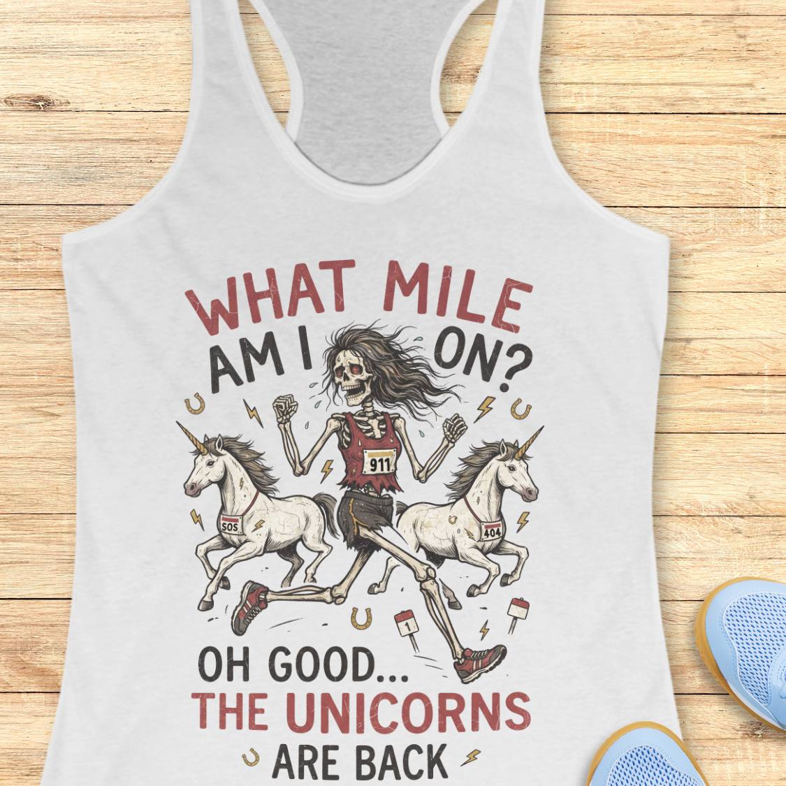 A white racerback tank top laid flat on a light wood surface, featuring a humorous graphic print of a skeleton runner racing two unicorns with the text 'WHAT MILE AM I ON? OH GOOD.