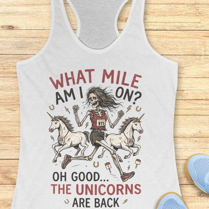 A white racerback tank top laid flat on a light wood surface, featuring a humorous graphic print of a skeleton runner racing two unicorns with the text 'WHAT MILE AM I ON? OH GOOD.