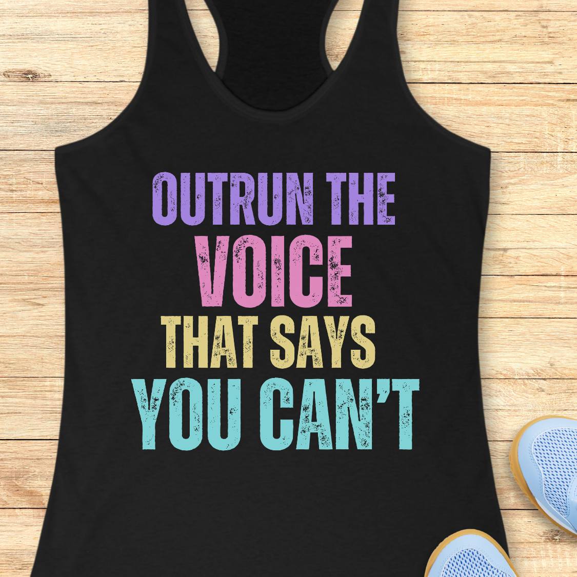A black racerback tank top laid flat on a light wood background, featuring the motivational text 'OUTRUN THE VOICE THAT SAYS YOU CAN'T' in purple, pink, yellow, and light blue distressed lettering. Partial light blue sneakers are visible at the bottom right as styling props.