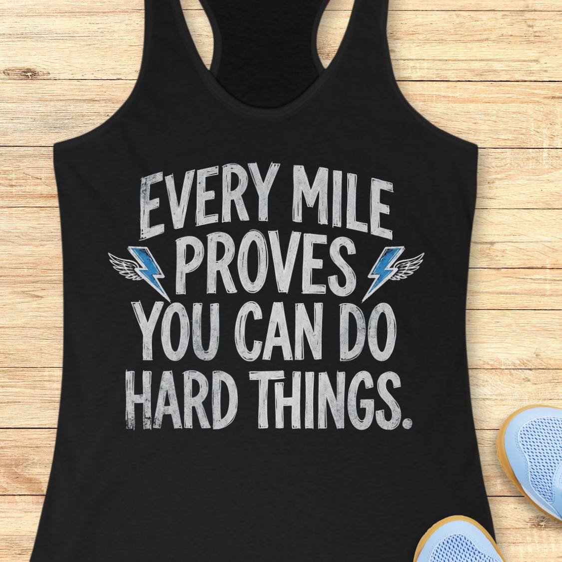 A black racerback tank top laid flat on a light wood surface, featuring the motivational text 'EVERY MILE PROVES YOU CAN DO HARD THINGS.' in distressed white lettering with blue winged lightning bolt accents.