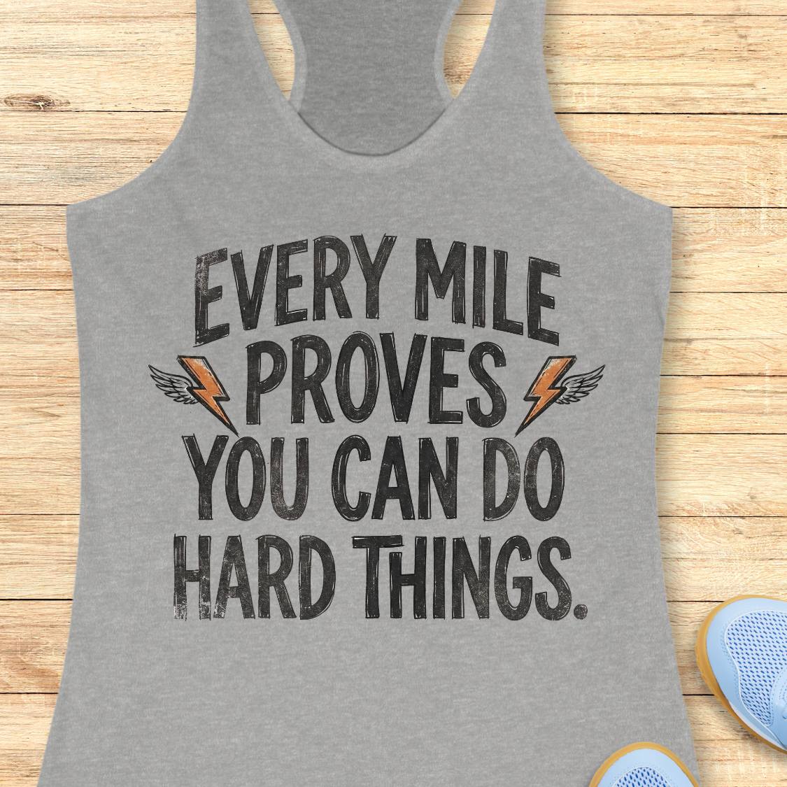 A heather gray racerback tank top with a motivational graphic reading 'EVERY MILE PROVES YOU CAN DO HARD THINGS.' in distressed black lettering, accented by two orange lightning bolts with wing details, laid flat on a light wood background.