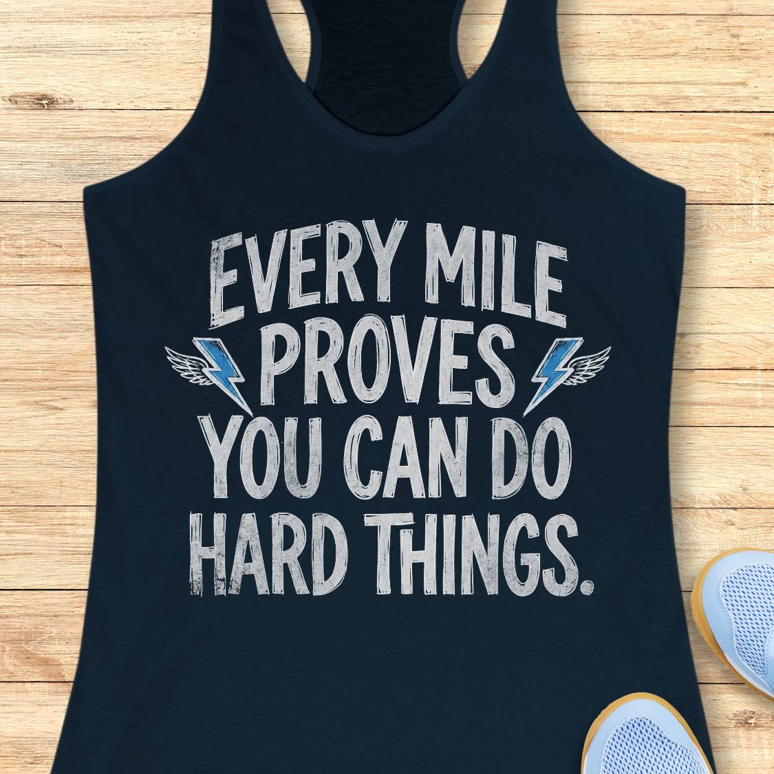 A navy blue racerback tank top with a motivational graphic reading 'EVERY MILE PROVES YOU CAN DO HARD THINGS.' in distressed white lettering, accented with two blue lightning bolts and small wing details, laid flat on a light wood background with partial sneakers visible at the edges.