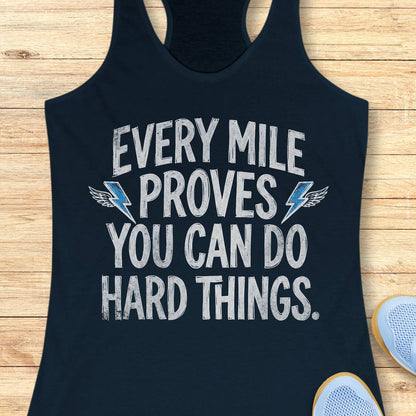 A navy blue racerback tank top with a motivational graphic reading 'EVERY MILE PROVES YOU CAN DO HARD THINGS.' in distressed white lettering, accented with two blue lightning bolts and small wing details, laid flat on a light wood background with partial sneakers visible at the edges.