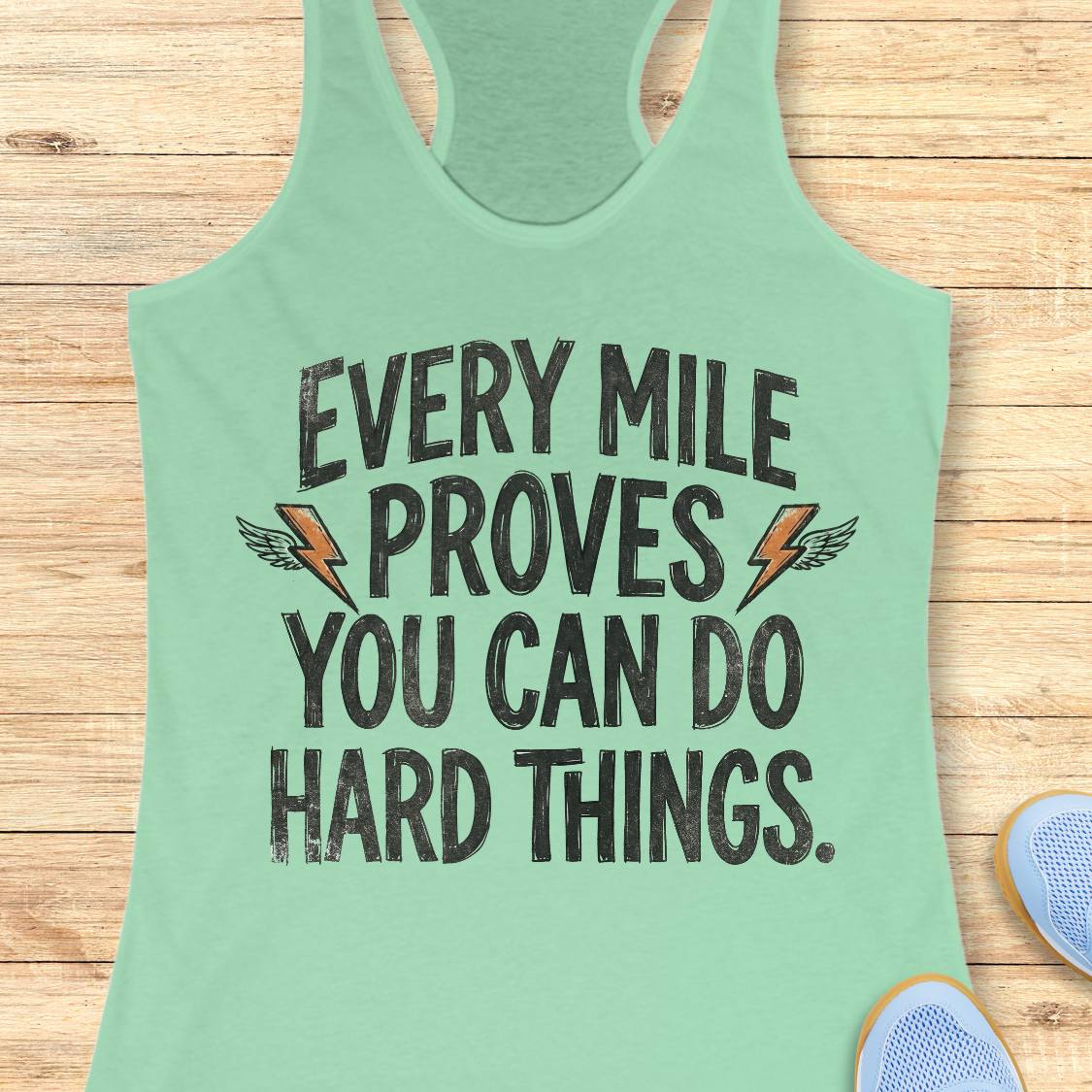 A mint green racerback tank top laid flat on a light wood background, featuring the motivational text 'EVERY MILE PROVES YOU CAN DO HARD THINGS.' in distressed black lettering with small orange lightning bolt and wing graphics.