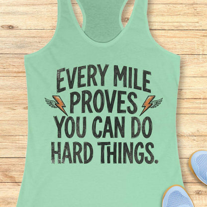 A mint green racerback tank top laid flat on a light wood background, featuring the motivational text 'EVERY MILE PROVES YOU CAN DO HARD THINGS.' in distressed black lettering with small orange lightning bolt and wing graphics.