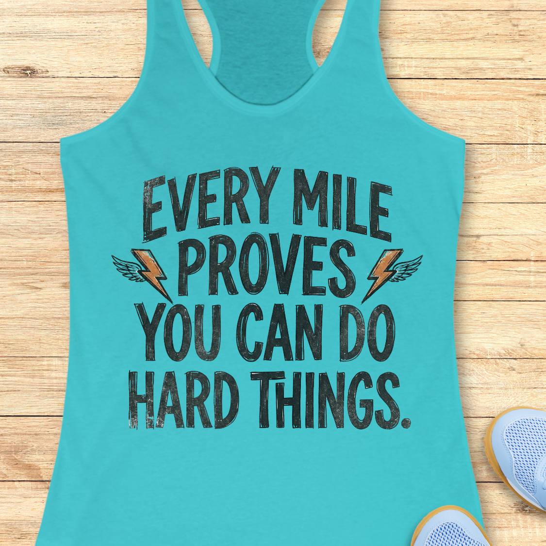 A turquoise racerback tank top laid flat on a light wood surface, featuring the motivational text 'EVERY MILE PROVES YOU CAN DO HARD THINGS.' in distressed black lettering with small orange lightning bolt and wing graphics.