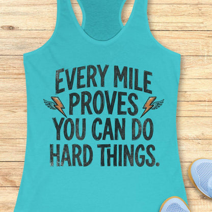 A turquoise racerback tank top laid flat on a light wood surface, featuring the motivational text 'EVERY MILE PROVES YOU CAN DO HARD THINGS.' in distressed black lettering with small orange lightning bolt and wing graphics.