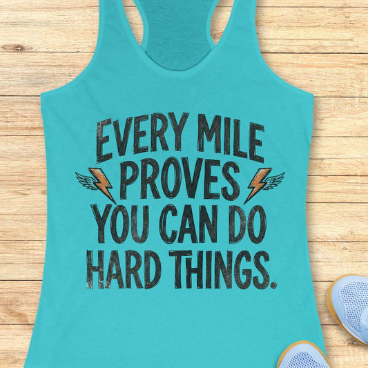 A turquoise racerback tank top laid flat on a light wood surface, featuring the motivational text 'EVERY MILE PROVES YOU CAN DO HARD THINGS.' in distressed black lettering with small orange lightning bolt and wing graphics.