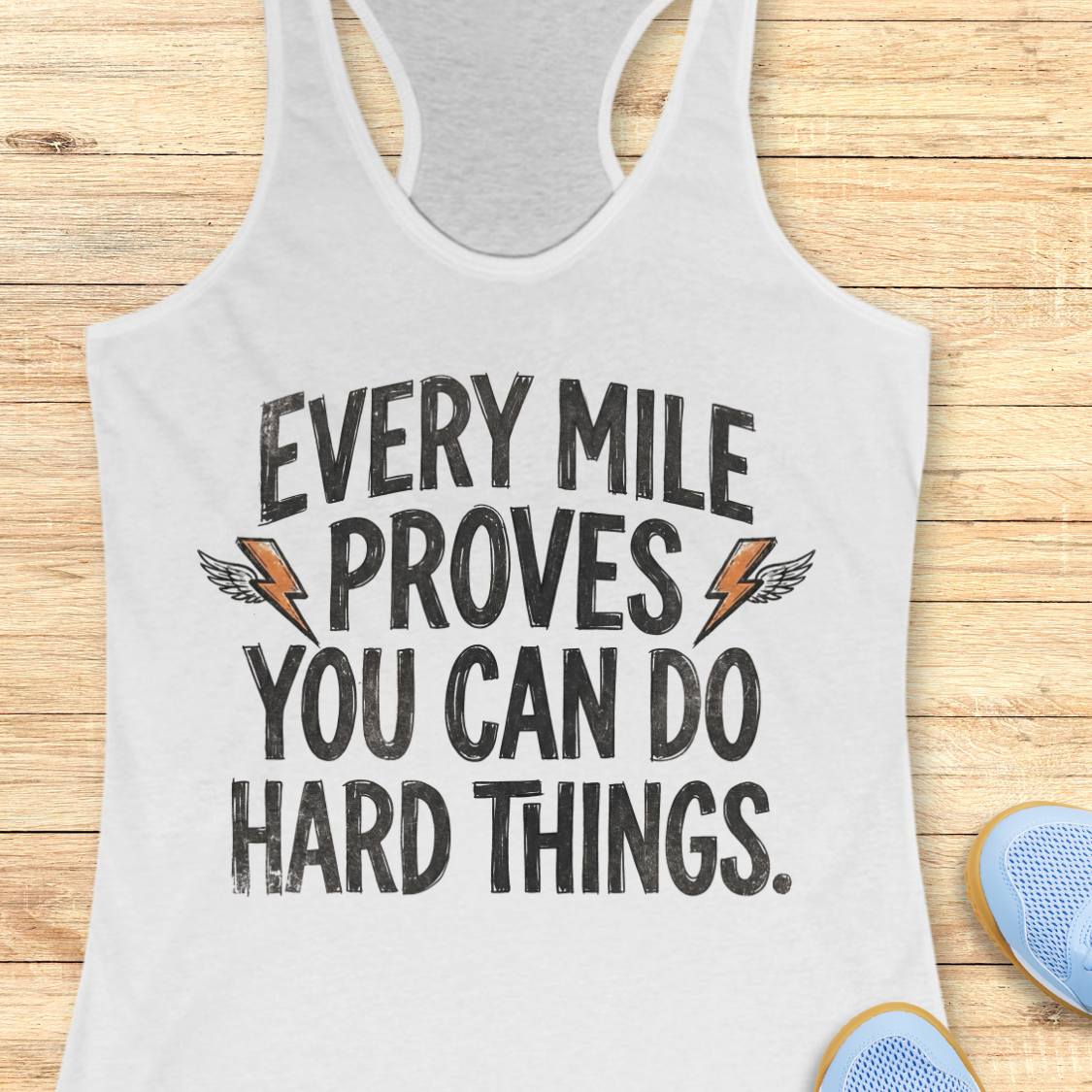 A white racerback tank top laid flat on a light wood plank background, featuring the motivational text 'EVERY MILE PROVES YOU CAN DO HARD THINGS.' in black distressed lettering with small orange lightning bolt and wing graphics.