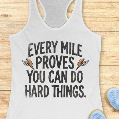 A white racerback tank top laid flat on a light wood plank background, featuring the motivational text 'EVERY MILE PROVES YOU CAN DO HARD THINGS.' in black distressed lettering with small orange lightning bolt and wing graphics.