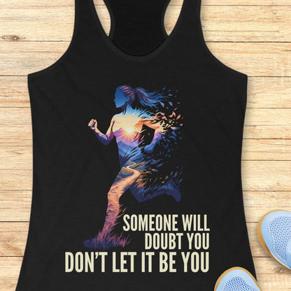 A black racerback tank top laid flat on a light wood surface, featuring a colorful graphic of a running woman with the text 'SOMEONE WILL DOUBT YOU DON'T LET IT BE YOU.' Part of a blue shoe is visible at the bottom right as a styling prop.
