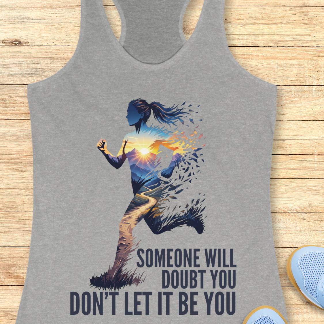 A heather gray racerback tank top featuring a graphic of a running woman with a colorful sunset inside her silhouette and the motivational text 'SOMEONE WILL DOUBT YOU DON'T LET IT BE YOU'. The tank top is displayed flat on a light wood plank background.