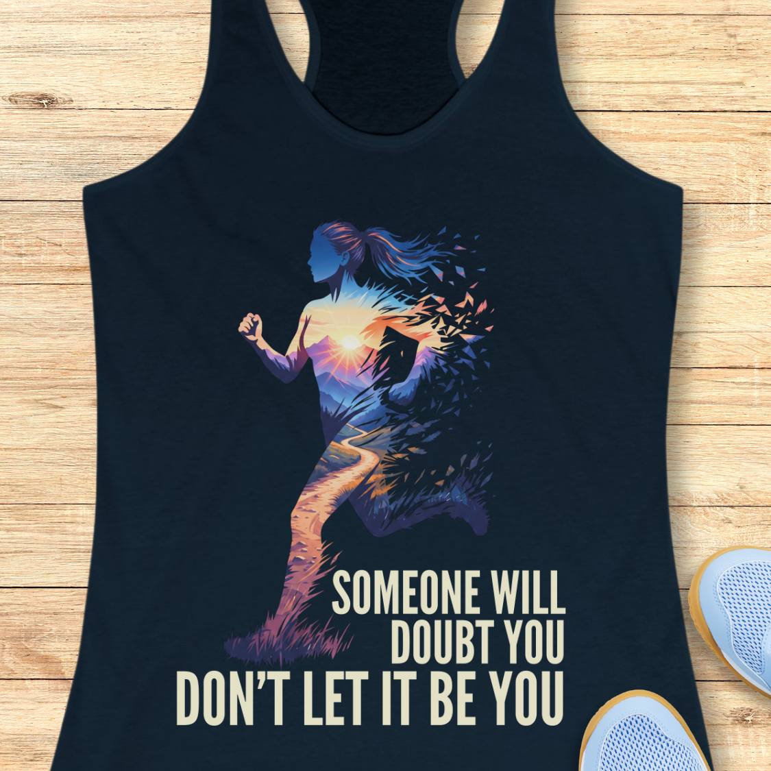 A navy racerback tank top laid flat on a light wood background, featuring a colorful graphic of a running woman silhouette with a sunset gradient and the text 'SOMEONE WILL DOUBT YOU DON'T LET IT BE YOU'. Partial white sneakers are visible at the bottom right as styling props.
