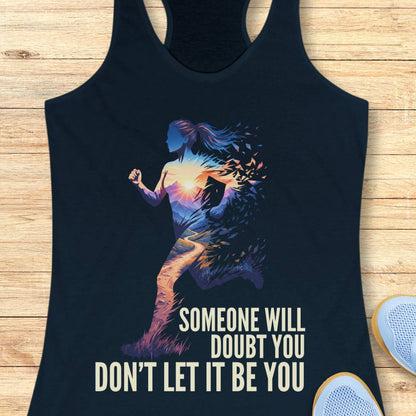 A navy racerback tank top laid flat on a light wood background, featuring a colorful graphic of a running woman silhouette with a sunset gradient and the text 'SOMEONE WILL DOUBT YOU DON'T LET IT BE YOU'. Partial white sneakers are visible at the bottom right as styling props.