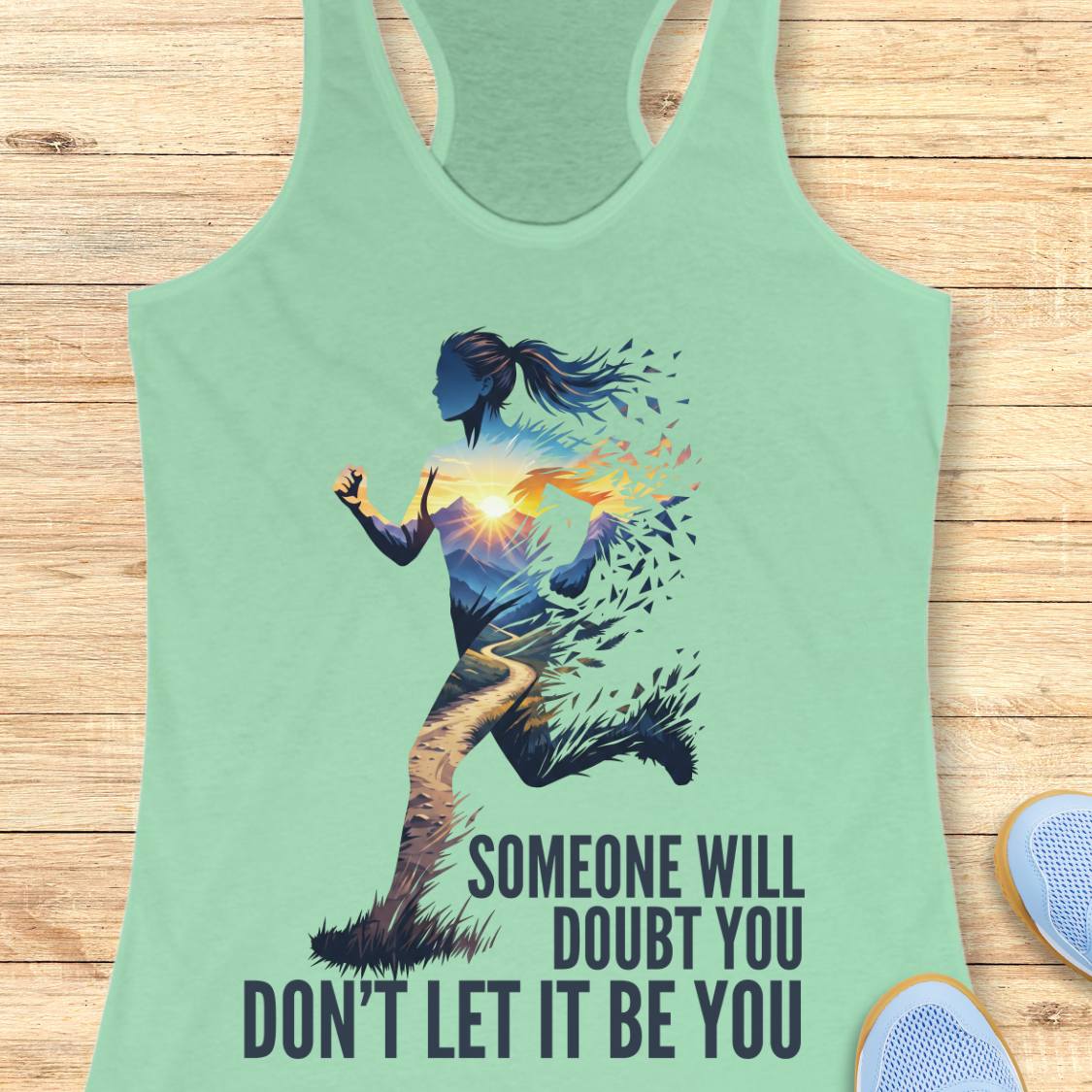 A mint green racerback tank top laid flat on a light wood background, featuring a graphic of a running female silhouette with a sunset and the text 'SOMEONE WILL DOUBT YOU DON'T LET IT BE YOU'.