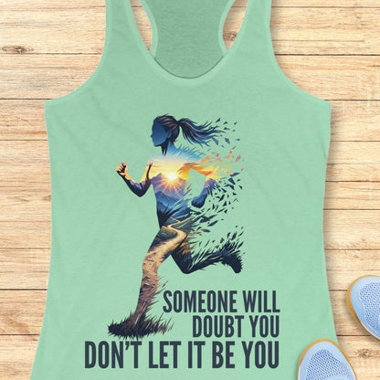 A mint green racerback tank top laid flat on a light wood background, featuring a graphic of a running female silhouette with a sunset and the text 'SOMEONE WILL DOUBT YOU DON'T LET IT BE YOU'.