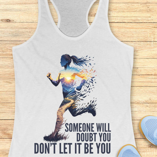 A white racerback tank top laid flat on a light wood background, featuring a graphic of a running female silhouette with a glowing sunburst and the text 'SOMEONE WILL DOUBT YOU DON'T LET IT BE YOU.' Partial blue sneakers are visible at the lower right as styling props.