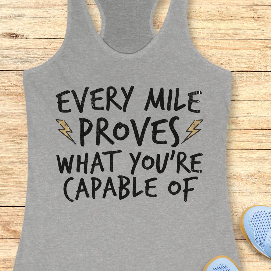 A heather gray racerback tank top with the motivational text 'EVERY MILE PROVES WHAT YOU'RE CAPABLE OF' in black lettering, accented with two small yellow lightning bolts, laid flat on a light wood background.