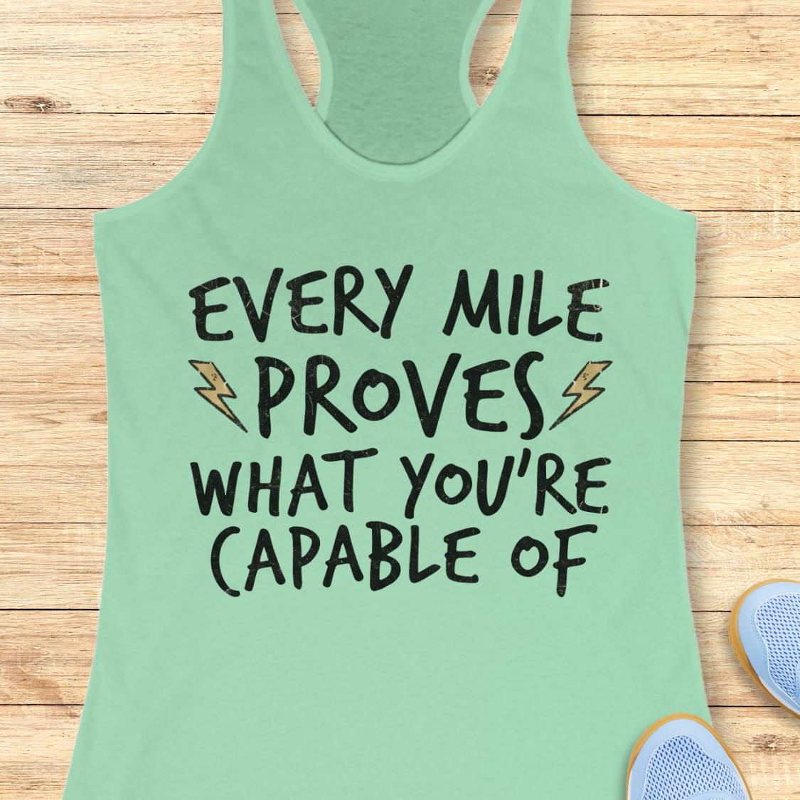 A mint green racerback tank top with the text 'EVERY MILE PROVES WHAT YOU'RE CAPABLE OF' in black lettering with small lightning bolt graphics, laid flat on a light wood background.