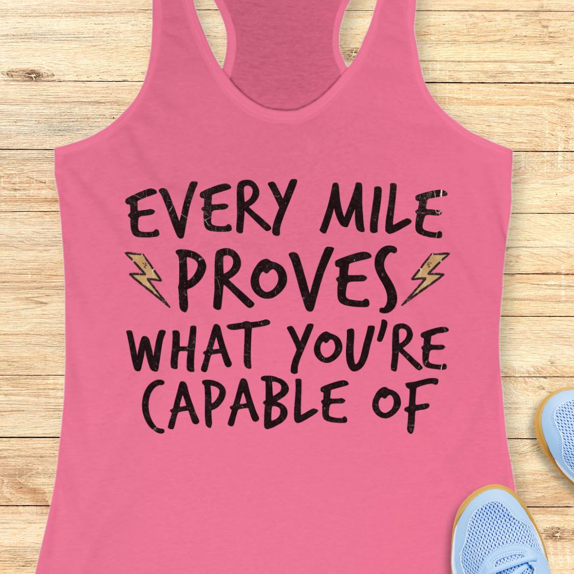 A pink racerback tank top laid flat on a light wood background, featuring the motivational text 'EVERY MILE PROVES WHAT YOU'RE CAPABLE OF' in black lettering with small yellow lightning bolt graphics. Partial blue sneakers are visible at the bottom corners as styling props.