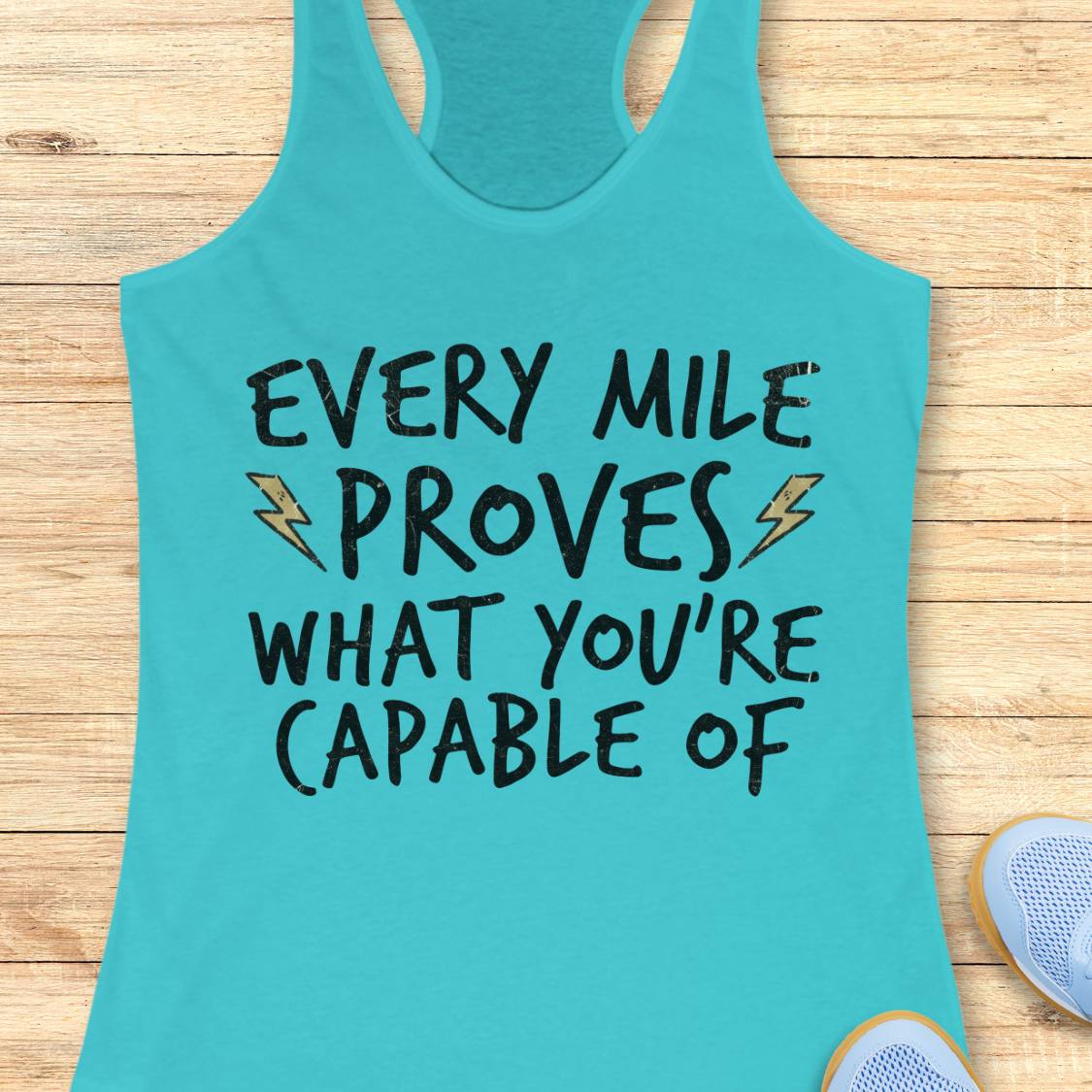A turquoise racerback tank top laid flat on a light wood background, featuring the motivational text 'EVERY MILE PROVES WHAT YOU'RE CAPABLE OF' in black lettering with small yellow lightning bolt accents. Partial shoes are visible at the bottom right as styling props.