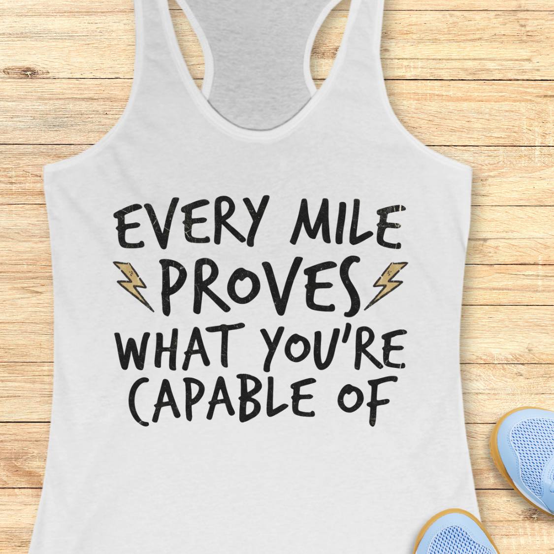 A white racerback tank top laid flat on a light wood background, printed with the motivational phrase 'EVERY MILE PROVES WHAT YOU'RE CAPABLE OF' in black lettering with small gold lightning bolt accents. Partial blue shoes are visible at the bottom right as styling props.